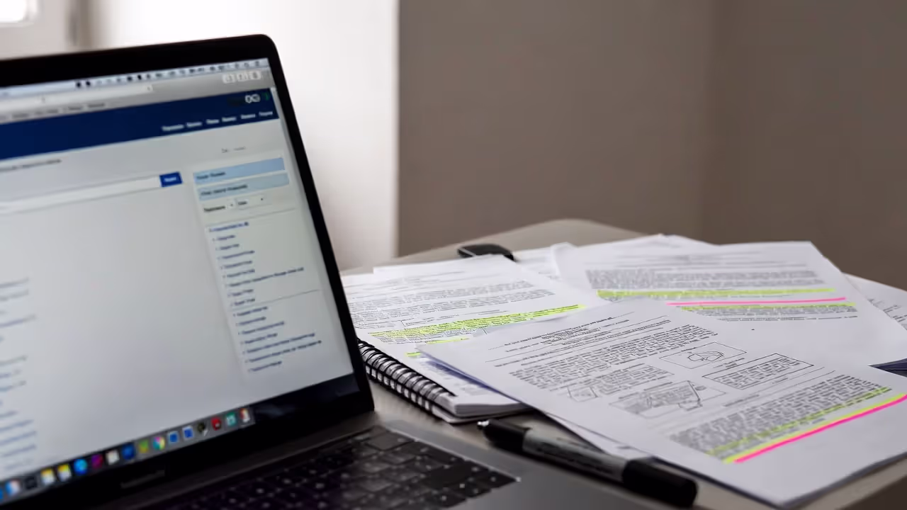 Laptop displaying a blurred patent database search with printed patent documents on desk